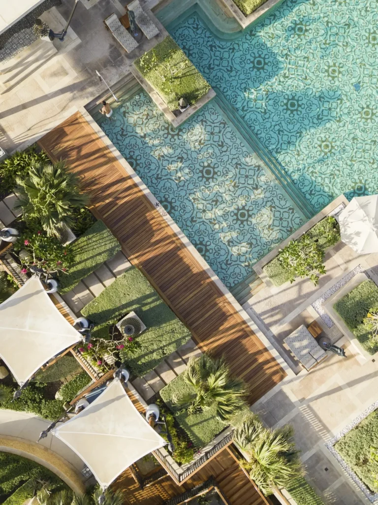 Aerial straight-down view of Jumeirah Al Naseem's pool revealing intricate Moroccan-style blue and white mosaic tile patterns in floral and geometric motifs, wooden deck walkway and tropical garden borders
