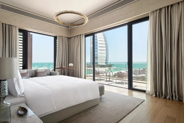Premier suite bedroom at Jumeirah Al Naseem with crisp king bed, brushed-gold ring chandelier and floor-to-ceiling glass doors framing a direct view of the Burj Al Arab and the Arabian Gulf