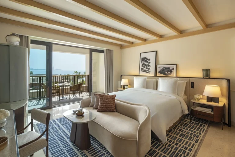 King bedroom at Jumeirah Al Naseem with white linen, exposed wood beam ceiling, sofa and floor lamp beside balcony doors open to rattan terrace furniture and a sea view