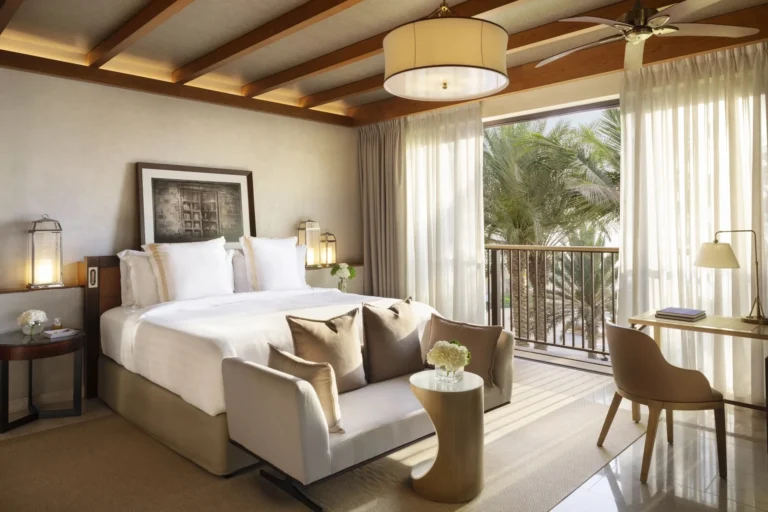 Arabian-style hotel bedroom at Jumeirah Al Naseem with wood beam and ceiling fan, ornate lantern bedside lamps, an antique-style headboard print, and balcony doors open to lush palm garden