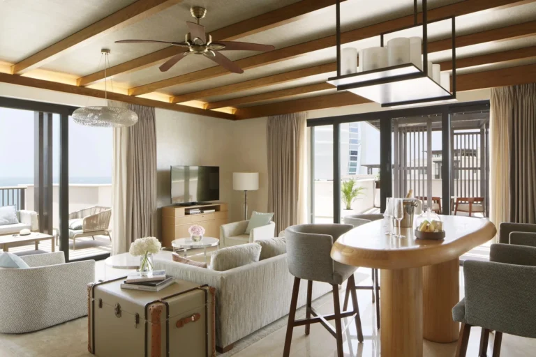 Penthouse-level suite bedroom at Jumeirah Al Naseem with ring chandelier, blue geometric carpet, arabesque lanterns on the nightstand, a chaise longue at the foot of the bed, and wide balcony doors to a furnished terrace