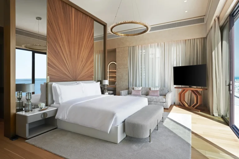 Contemporary suite bedroom at Jumeirah Al Naseem with a sculptural fan-pattern wood headboard wall, ring chandelier, pale grey carpet, bench at the foot and floor-to-ceiling windows with sea views