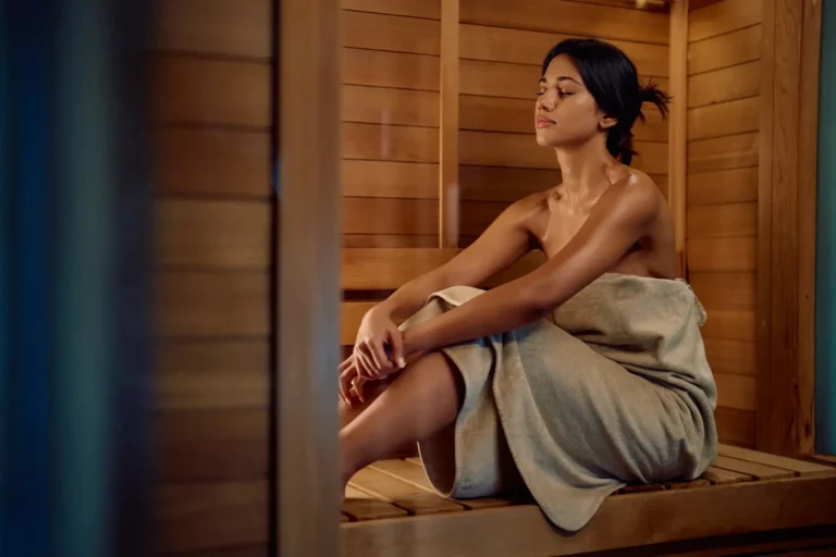 Woman wrapped in a grey towel sitting alone in a wood-panelled cedar sauna at Jumeirah Al Naseem's Talise Spa, eyes closed in quiet relaxation