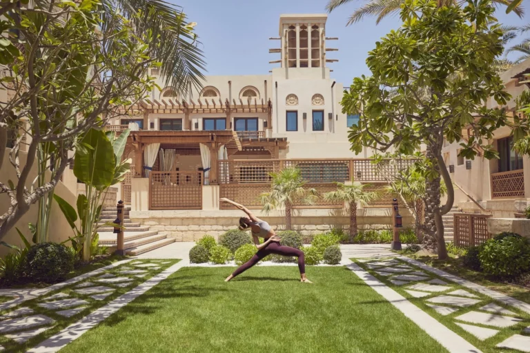 Woman in yoga warrior pose on the manicured lawn of Jumeirah Al Naseem's garden courtyard with the hotel's ornate Arabic windtower facade, arched pergolas and frangipani trees behind her