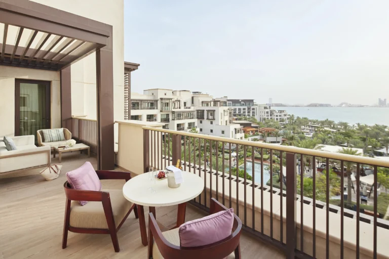 Private room terrace at Jumeirah Al Naseem with two rattan chairs and a table set with champagne and a fruit bowl, looking over the resort's gardens and free-form pool to the Arabian Gulf and Palm Jumeirah on the horizon