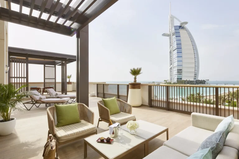 Expansive suite terrace at Jumeirah Al Naseem with a pergola, sun loungers, rattan seating and a coffee table set with champagne, looking directly at the Burj Al Arab and the curved wave-facade of the Jumeirah Beach Hotel beyond the resort gardens