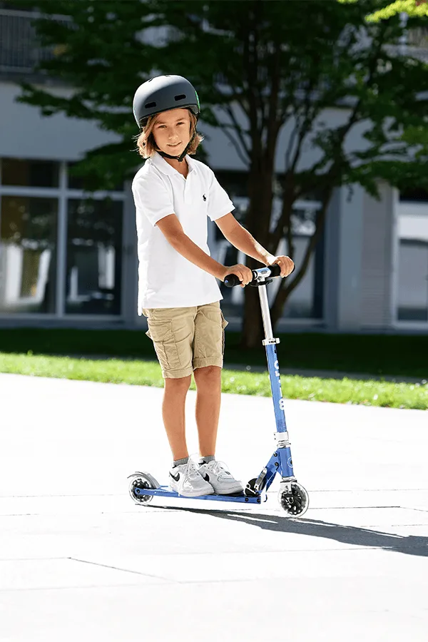 Child riding Micro Sprite LED Blue Stripe scooter outdoors wearing a helmet