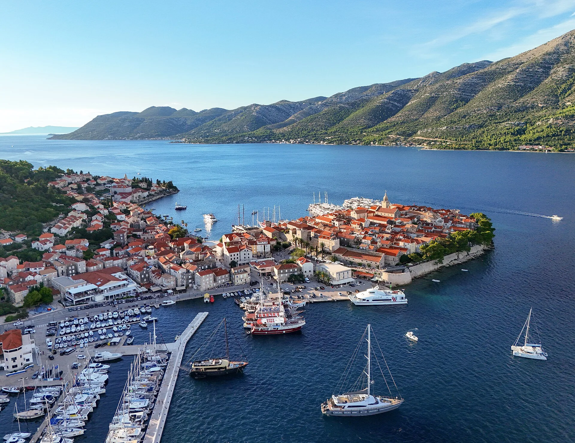 Aerial view of Korčula's walled Old Town on its peninsula, with sailing yachts moored in the marina and hills beyond