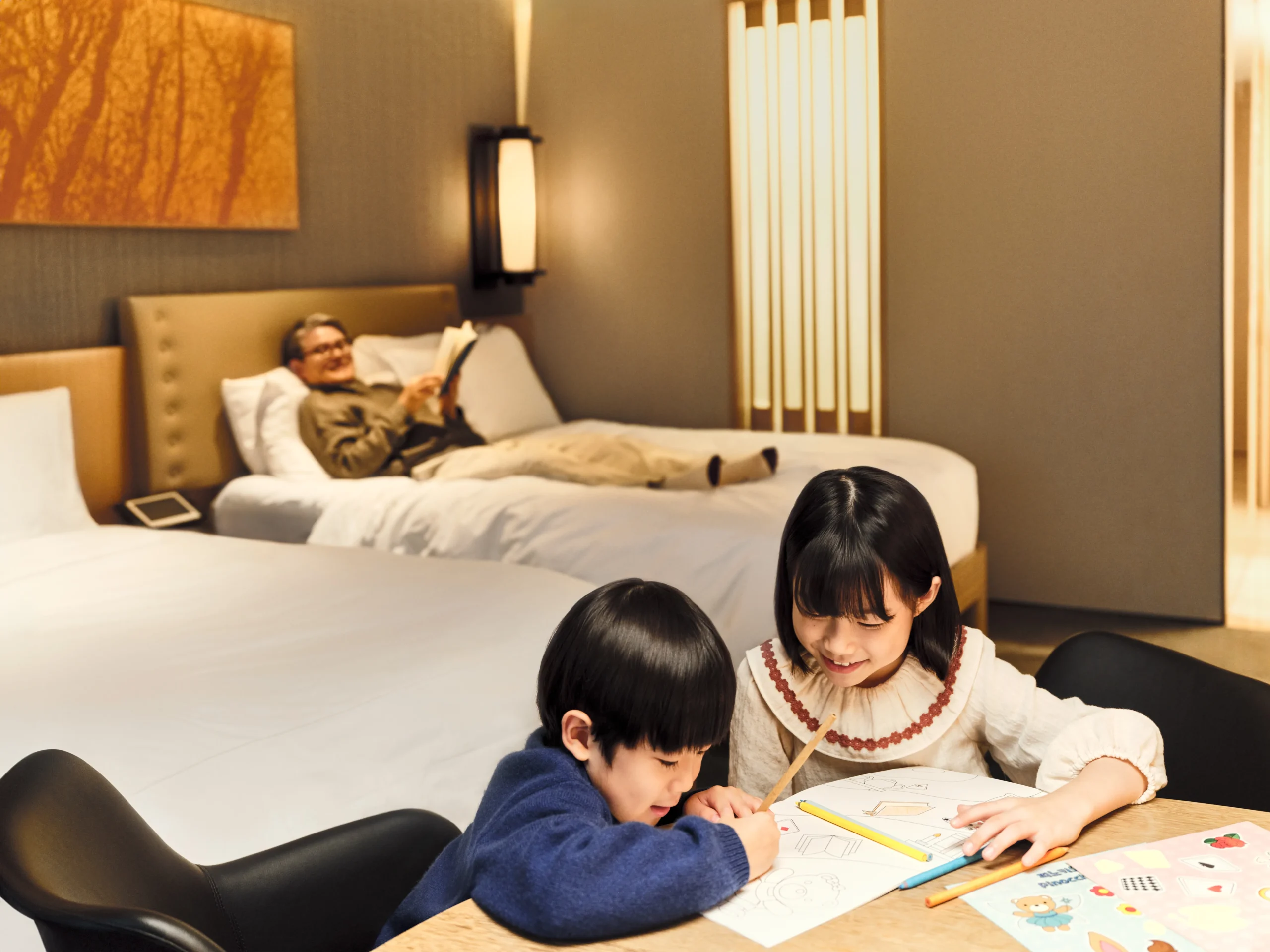 A grandfather reads on a hotel bed while two grandchildren color at a nearby desk in the same spacious modern guest room.