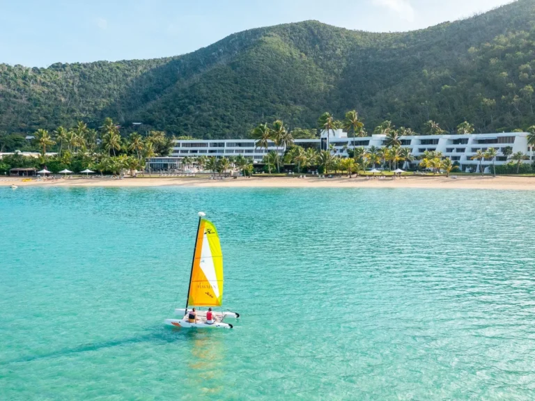 A small sailing catamaran on turquoise water in front of the beachfront resort wing and green hills