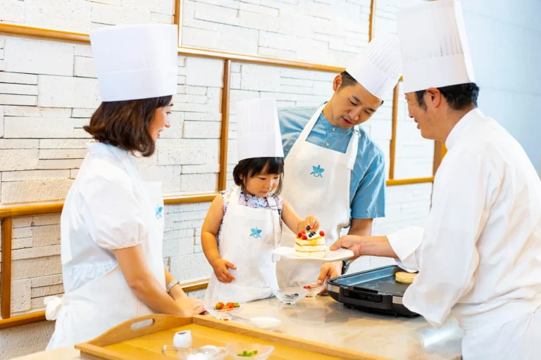 A small girl in a chef's hat and Halekulani apron tops a stack of pancakes with berries while two adults and a chef in whites look on