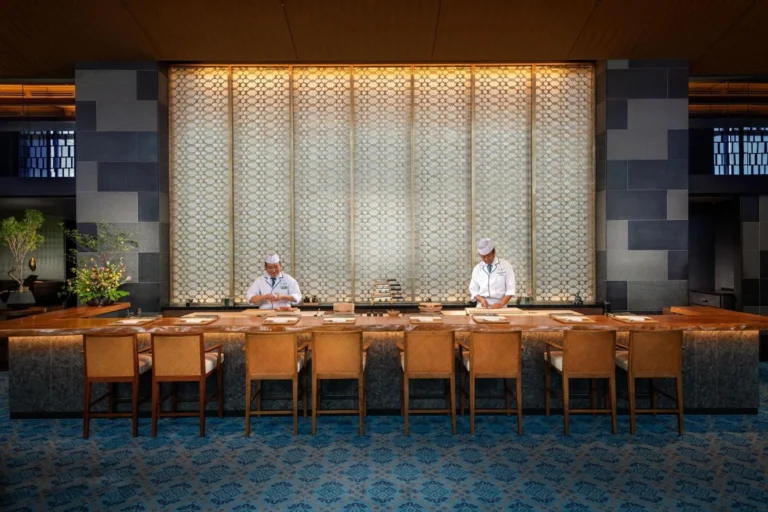 Sushi chefs behind a wooden counter with tan chairs and a lit screen wall