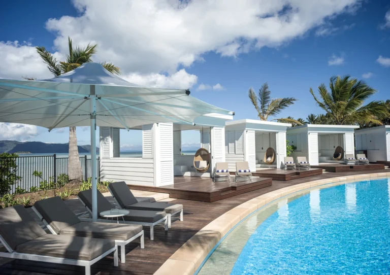 White poolside cabanas and sun loungers beside the curved pool with the sea beyond