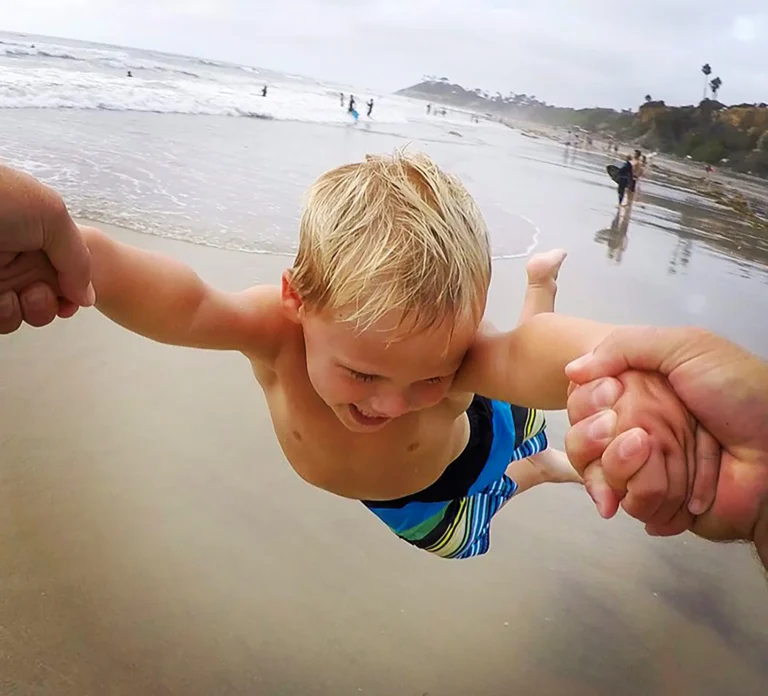 A laughing toddler is swung through the shallows of a beach by two adults holding his hands.