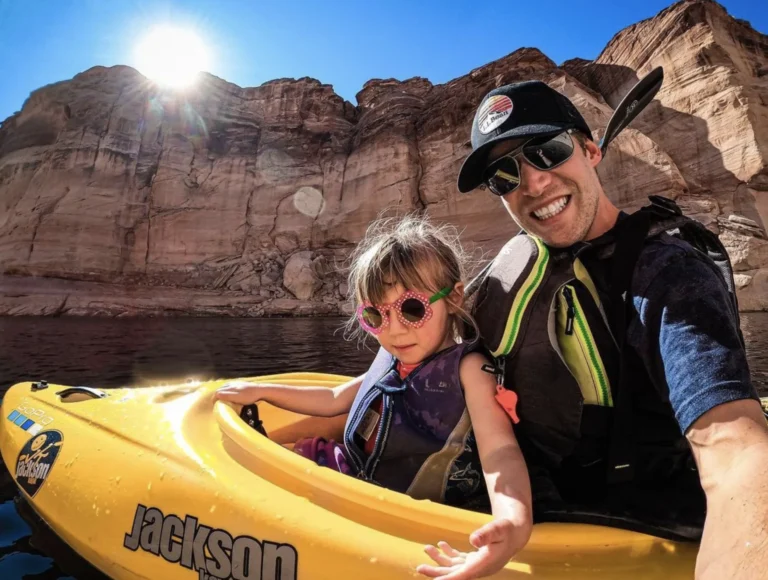 A father and his young daughter, both in life jackets, share a yellow kayak below red sandstone cliffs.