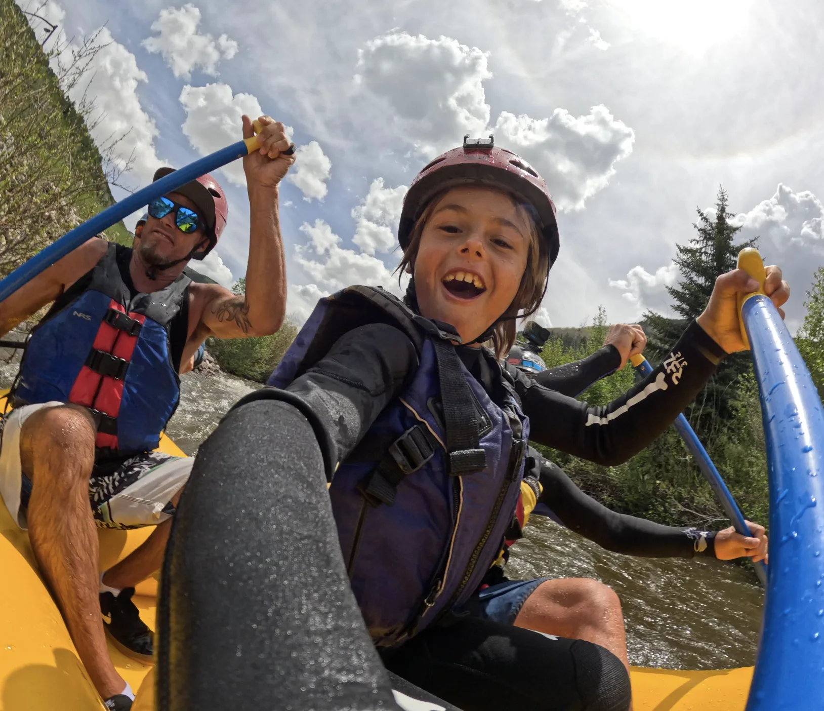 A smiling boy in a helmet and life jacket sits in a yellow raft beside an adult on a river.