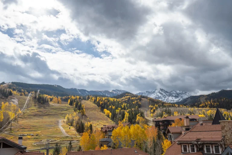 Panoramic view over Mountain Village rooftops, autumn aspens and a green ski run below distant peaks