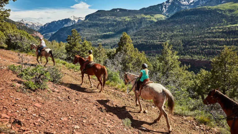 Riders on horses descend a rocky trail with a green valley and mountains in the background, two of them children