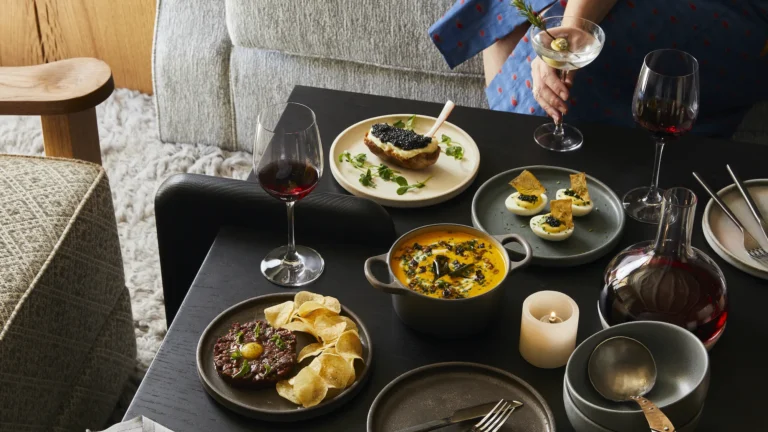 Dark table with small plates, a bowl of soup, wine glasses and a martini beside a soft sofa