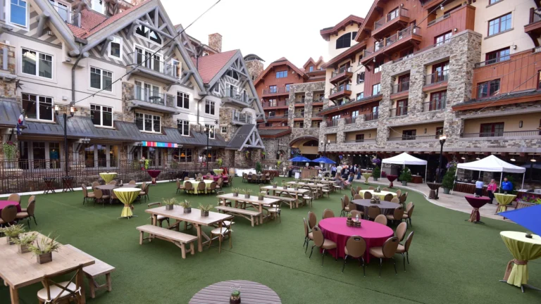 Hotel plaza with a green lawn, picnic tables and umbrellas surrounded by the gabled timber-and-stone building