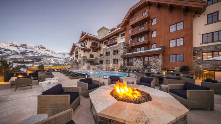 Stone fire pit with wicker armchairs on the pool terrace at dusk, steaming pool and snowy peaks behind the timber facade