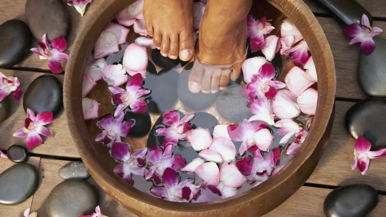 Bare feet in a wooden bowl of pink flower petals among dark smooth stones