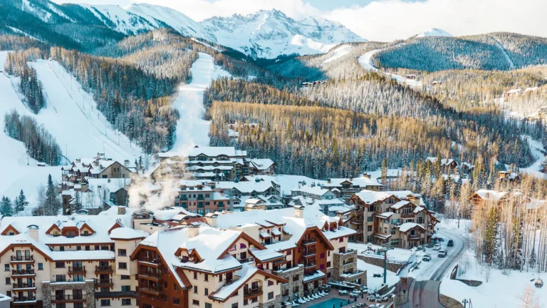 Aerial winter view of Mountain Village rooftops with a groomed ski run and forested slopes behind