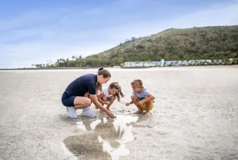 An adult in a navy uniform and two children crouching over a tide pool on the sand