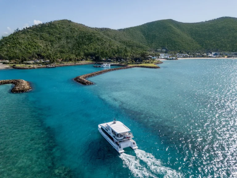 A catamaran leaving the marina breakwater in clear turquoise water below green hills