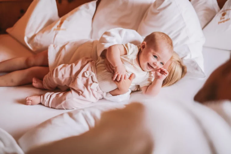Smiling baby lying on white hotel bedding with a parent's arm and legs beside it