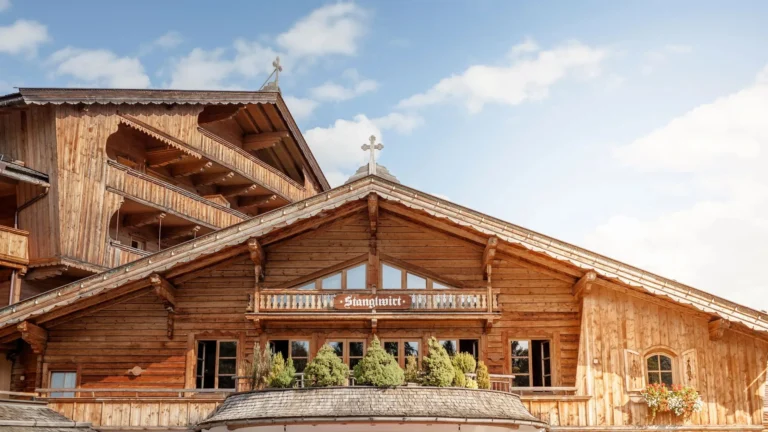 Weathered timber facade of the main house with carved balcony, gabled roof and the Stanglwirt sign under a blue sky
