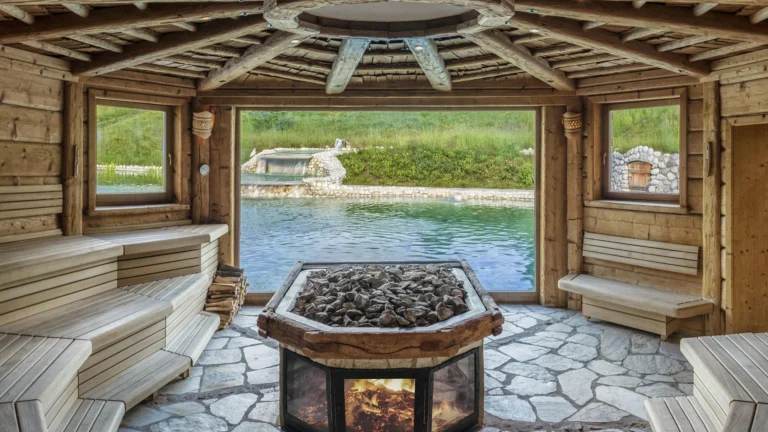 Round timber sauna with a central fire pit, tiered benches and a wide window onto a natural bathing pond