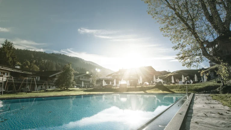 Outdoor pool at sunrise with a bright sun flare, trees, swings and resort buildings behind