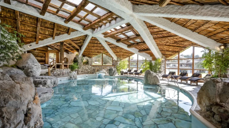 Indoor pool with rock formations and flagstone floor beneath a timber-beam skylight roof, loungers along the glass wall