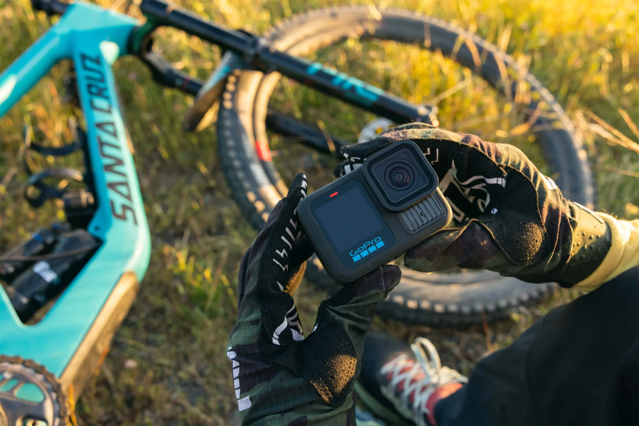 A gloved hand holds a GoPro camera beside a turquoise mountain bike lying in the grass.