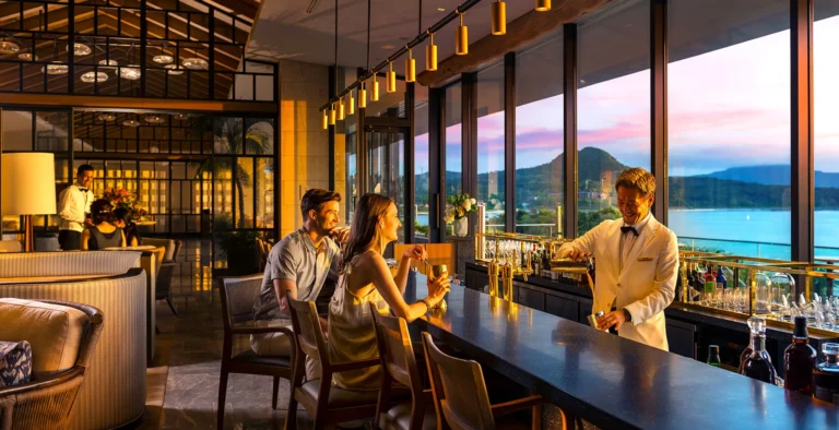 Two guests at a bar counter served by a bartender in a white jacket with a sunset sea view behind
