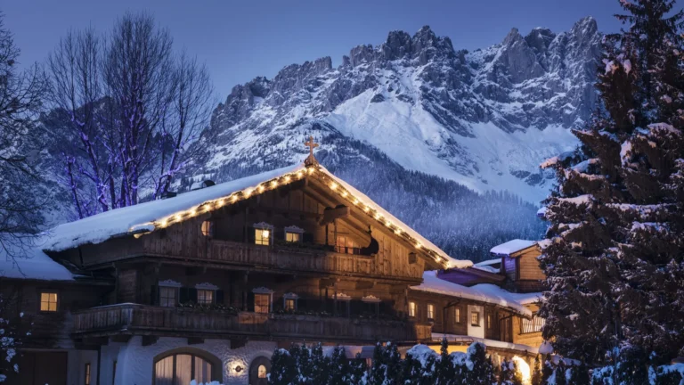Snow-covered timber chalet with warm string lights at dusk and the snowy Wilder Kaiser peaks behind