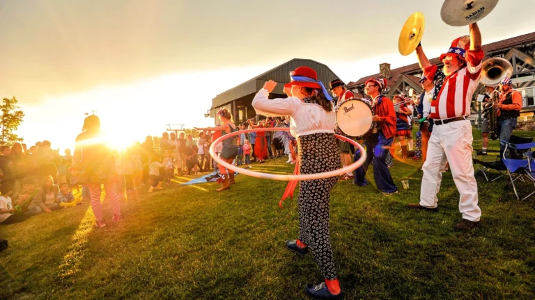 Young children sit and watch on a lawn at sunset as a costumed brass band plays and a performer spins a hula hoop