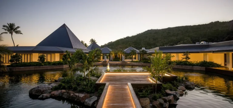 Timber boardwalk crossing a lily pond to a pyramid-roofed pavilion at dusk
