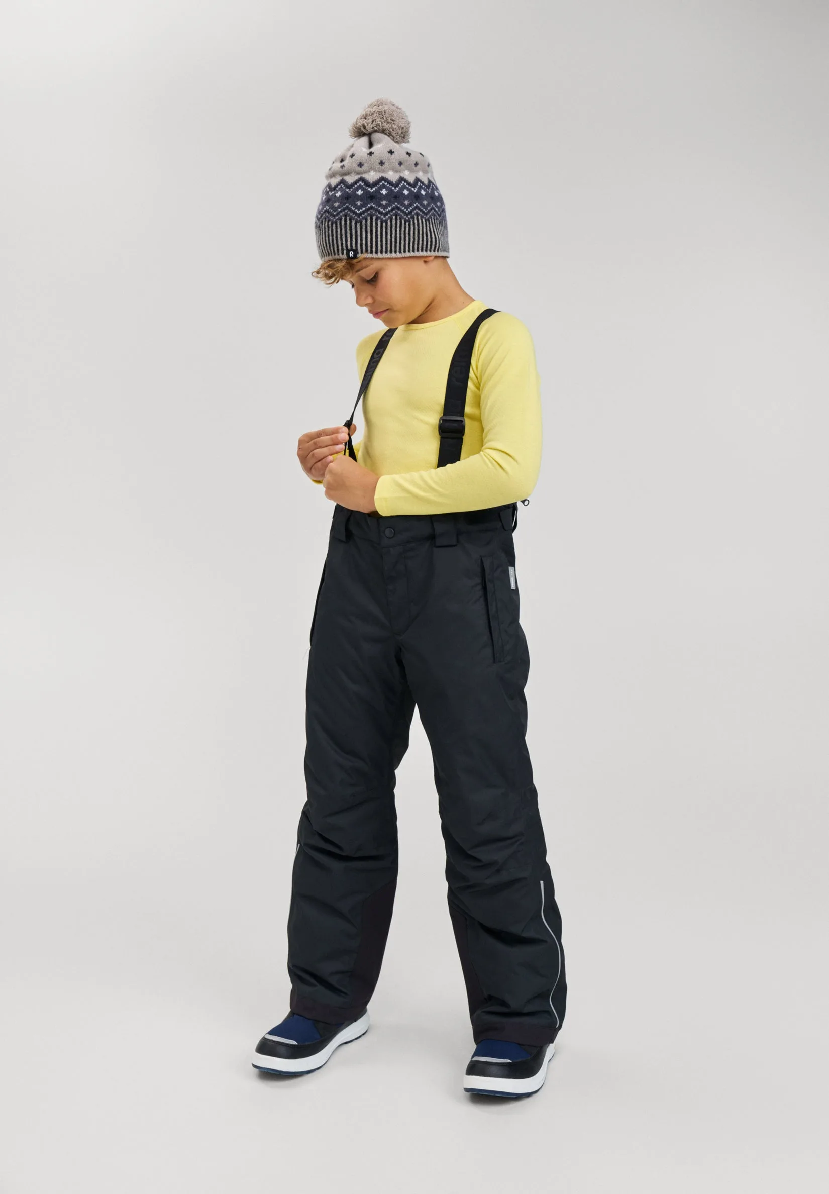 A boy in black Reima Wingon ski pants with the suspenders over his shoulders, a yellow top and a patterned bobble hat, standing against a light grey studio backdrop