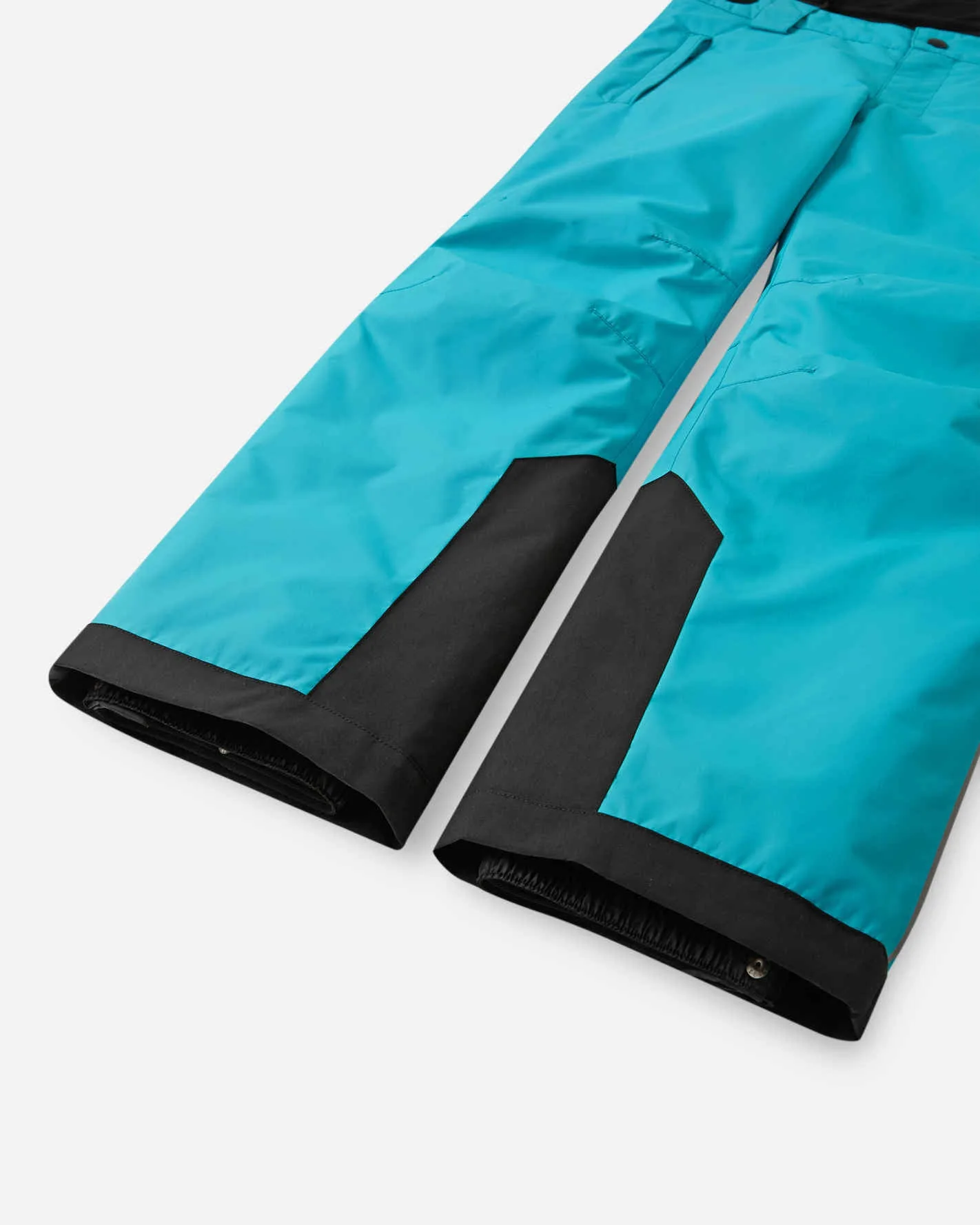 Both leg ends of turquoise Reima Wingon ski pants, each with a black reinforcement panel and an inner snow gaiter with a snap fastener, on a white background