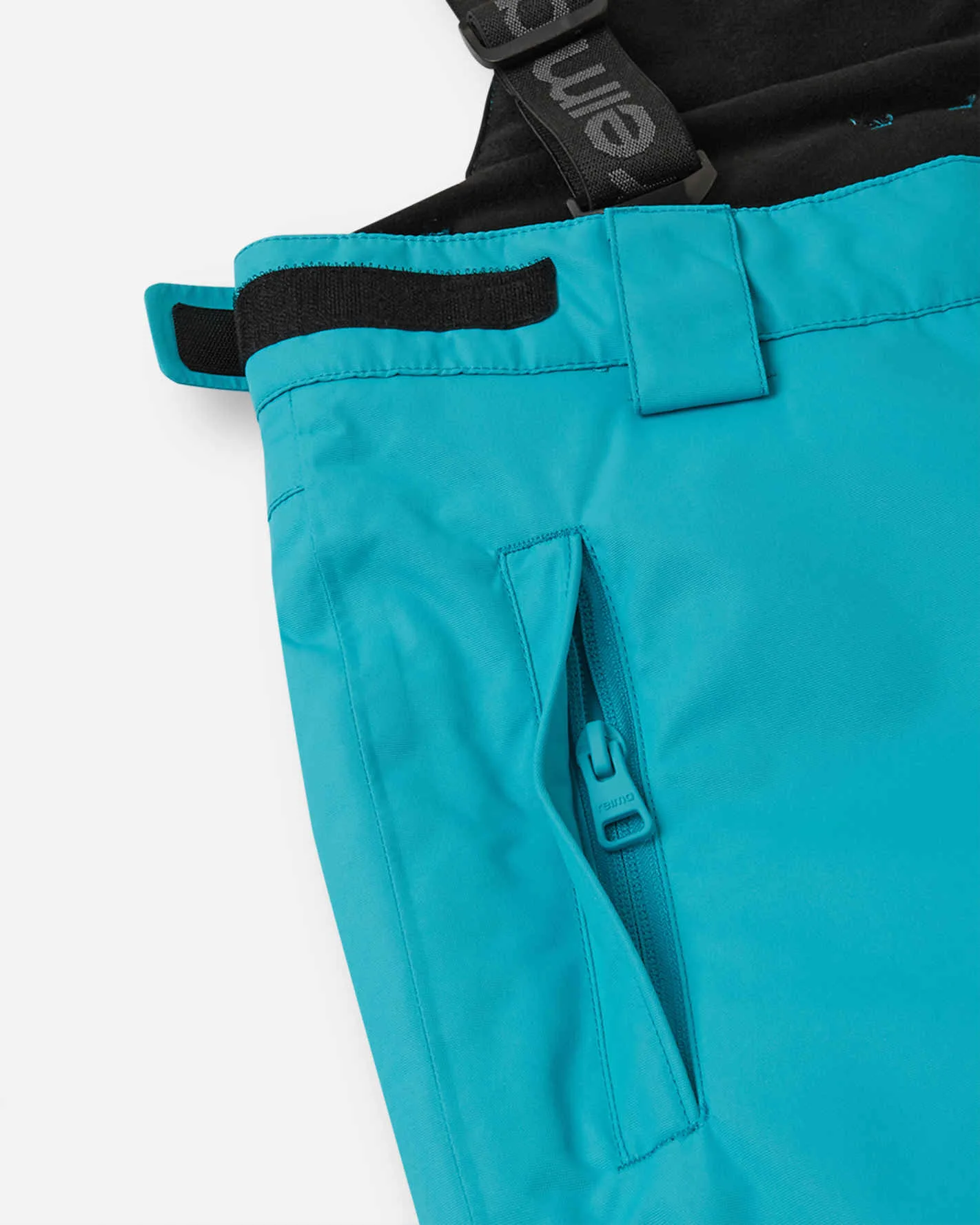 Close-up of the waistband of turquoise Reima Wingon ski pants with a black velcro adjuster tab, a belt loop, a side zip pocket and a suspender strap printed with the Reima name