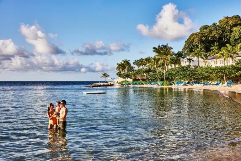 Parents standing in knee-deep sea water holding a toddler, with the beach and palms behind