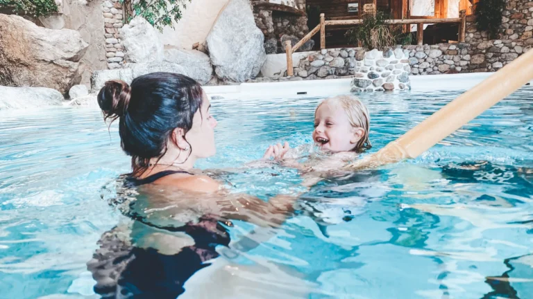 Woman helping a laughing child swim with a foam noodle in a pool surrounded by stone walls