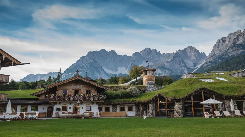 Timber farmhouse with a flower balcony and a grass-roofed spa building below the jagged Wilder Kaiser range under a blue sky