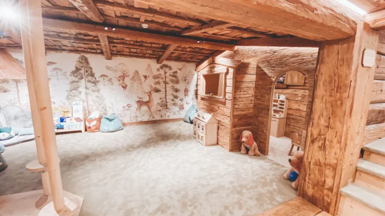 Children's play room with a sandy floor, a mural of forest animals, a timber play house and soft toys