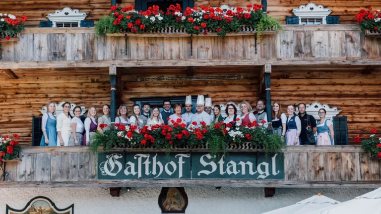 Hotel team in dirndl, aprons and chef whites standing on the flower-decked balcony of Gasthof Stangl